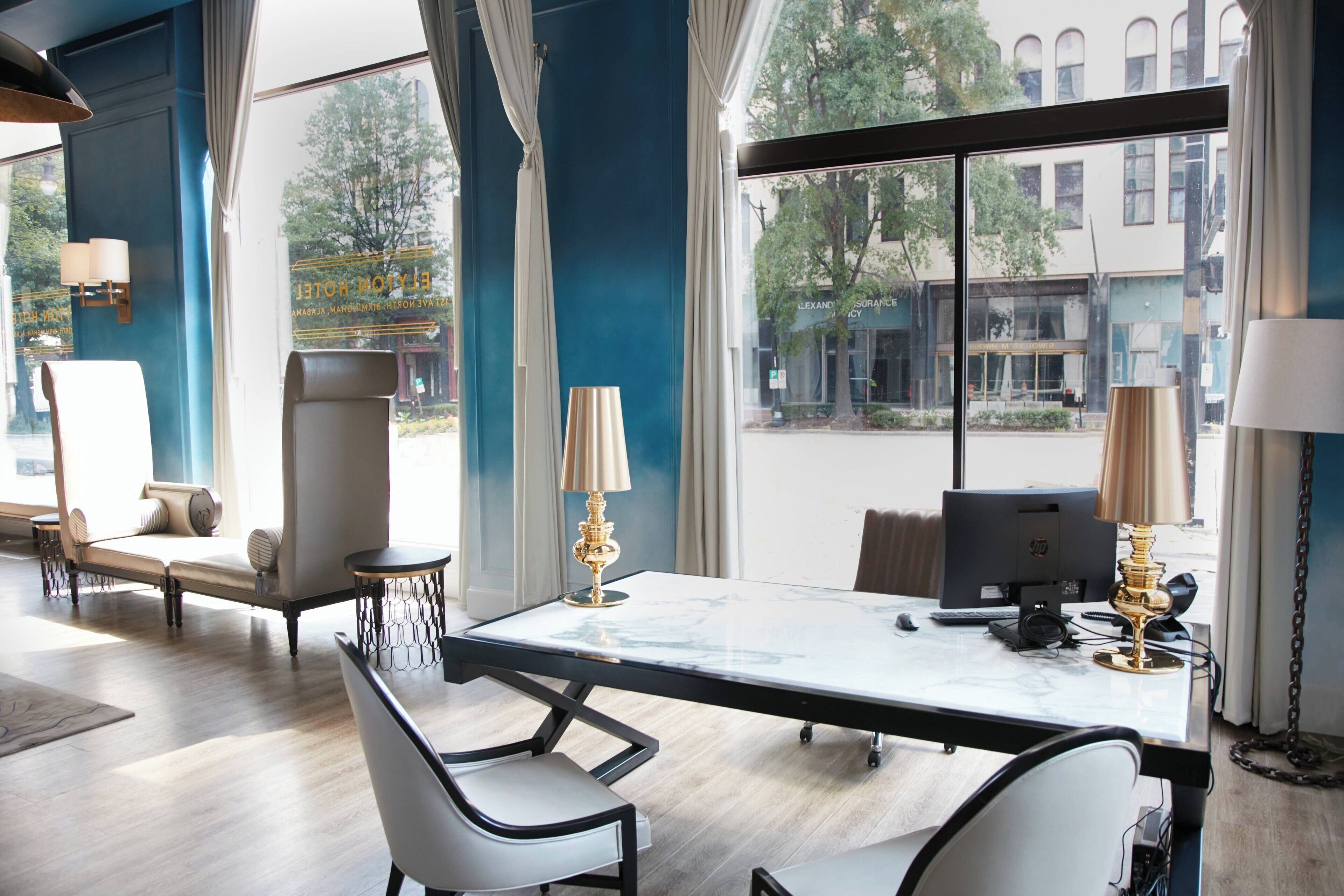 Una habitación de hotel moderna con un ventanal grande, un escritorio de mármol, dos sillas, una computadora y una lámpara.