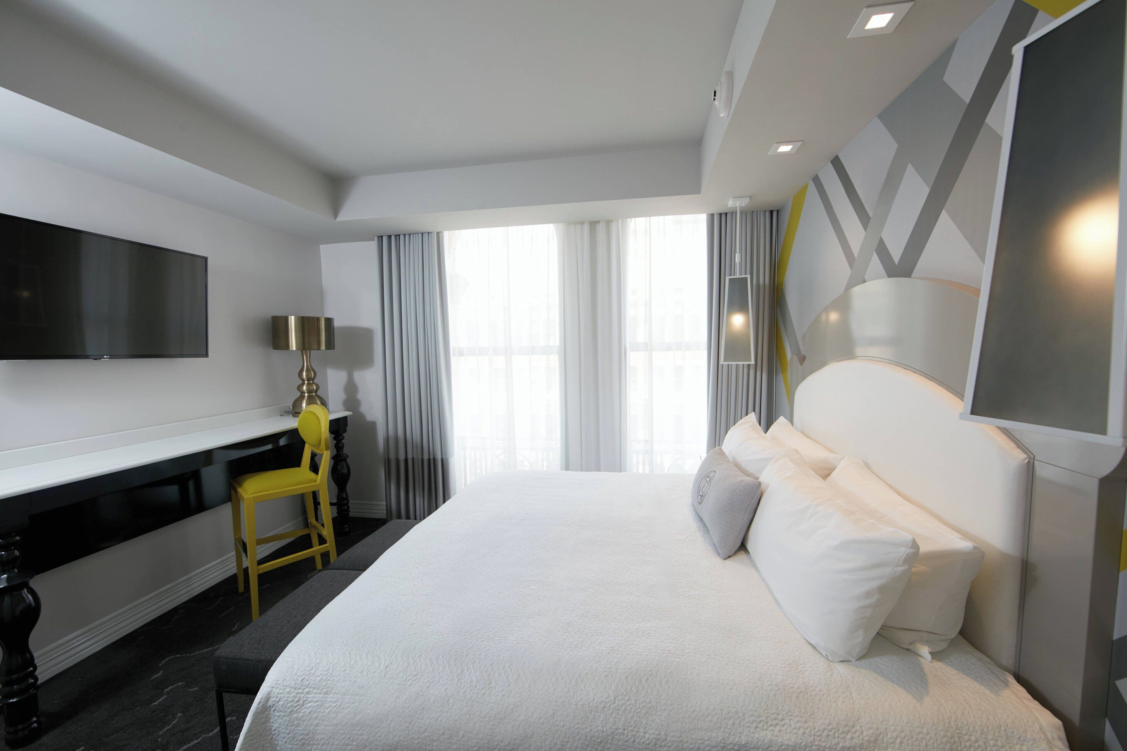 Una habitación de hotel moderna con una cama grande, un escritorio, un televisor de pantalla plana y un ventanal con cortinas.