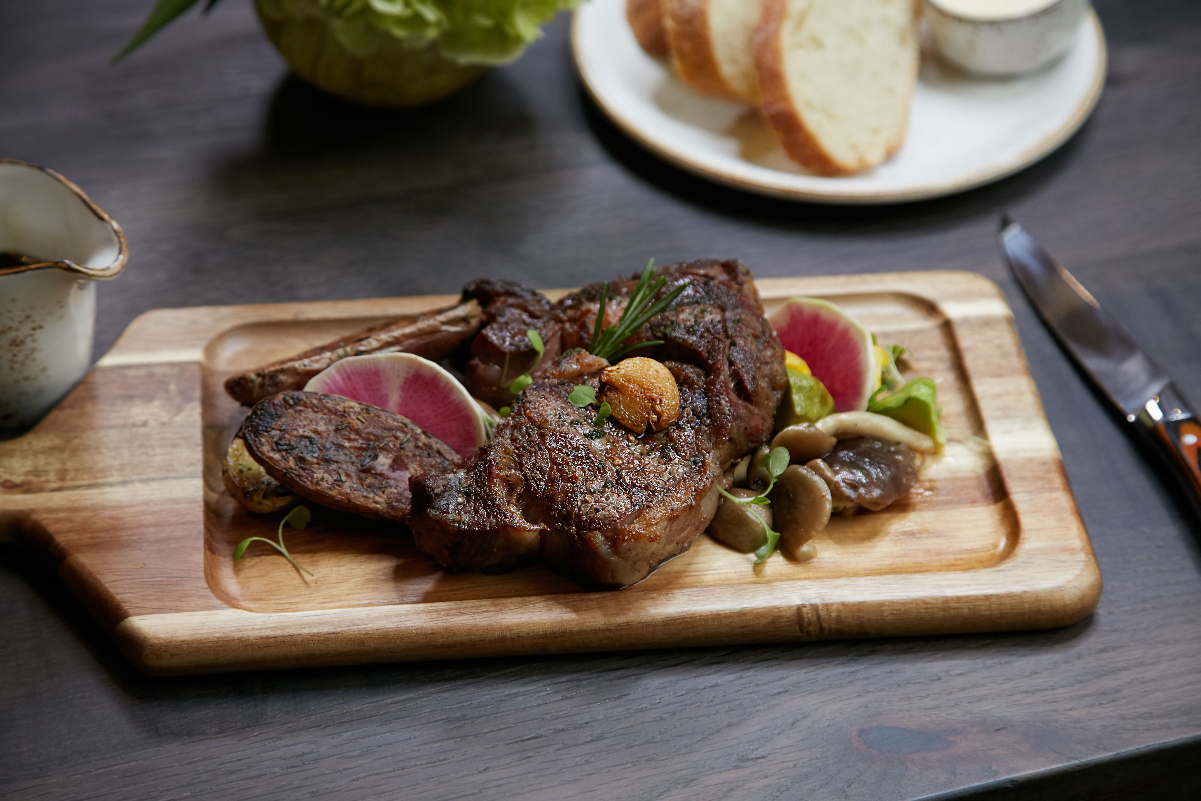 Una tabla de madera con un bistec, champiñones y vegetales de hoja verde sobre una mesa oscura.