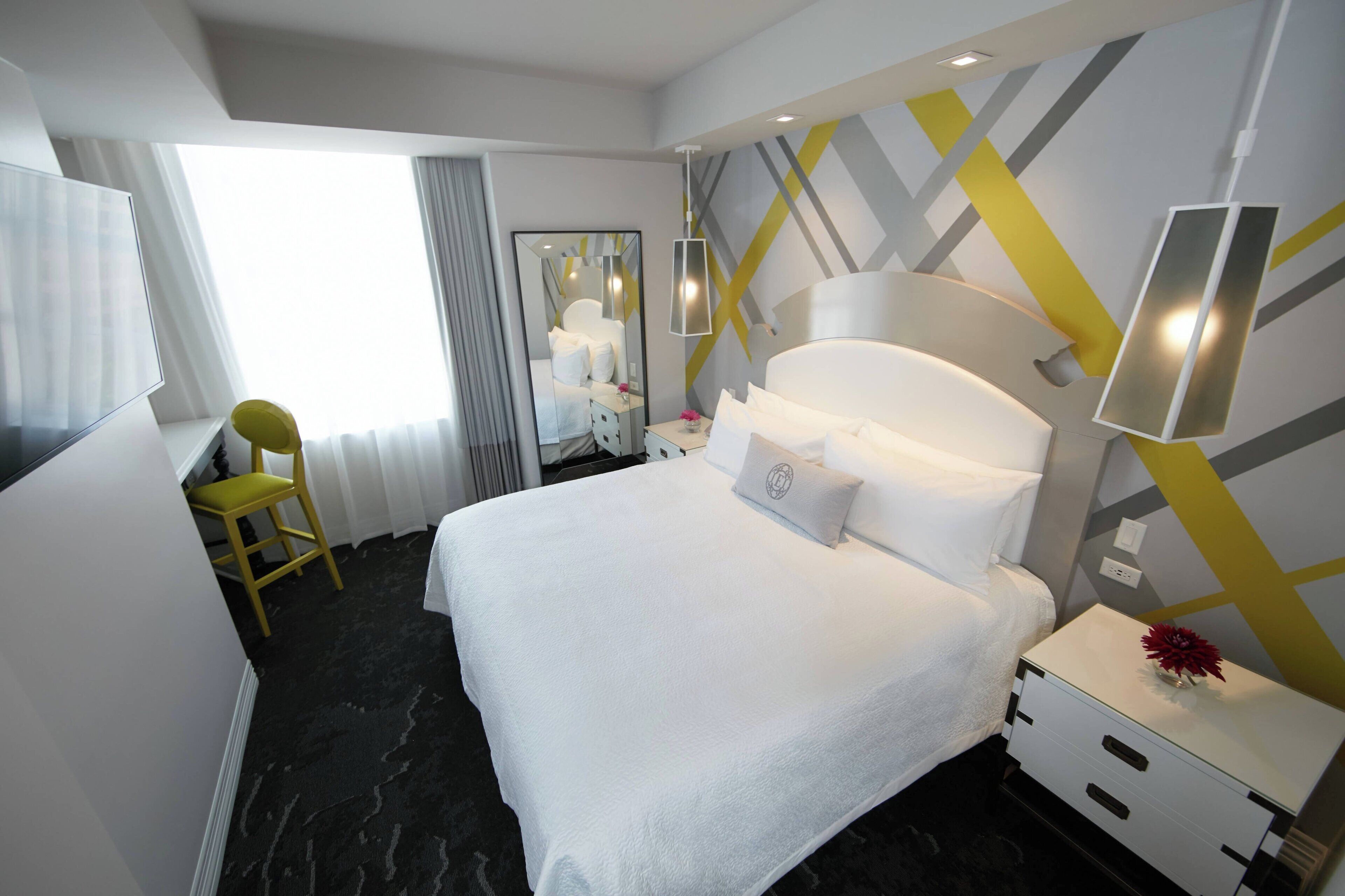 A hotel room with a large bed, a bedside table with a lamp, a mirror, a window with curtains, and a yellow chair.