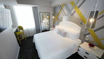 A hotel room with a large bed, a bedside table with a lamp, a mirror, a window with curtains, and a yellow chair.