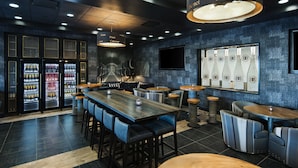 A restaurant interior with a dark blue tiled floor, wooden tables and chairs, a wine cooler, and a wall-mounted TV.