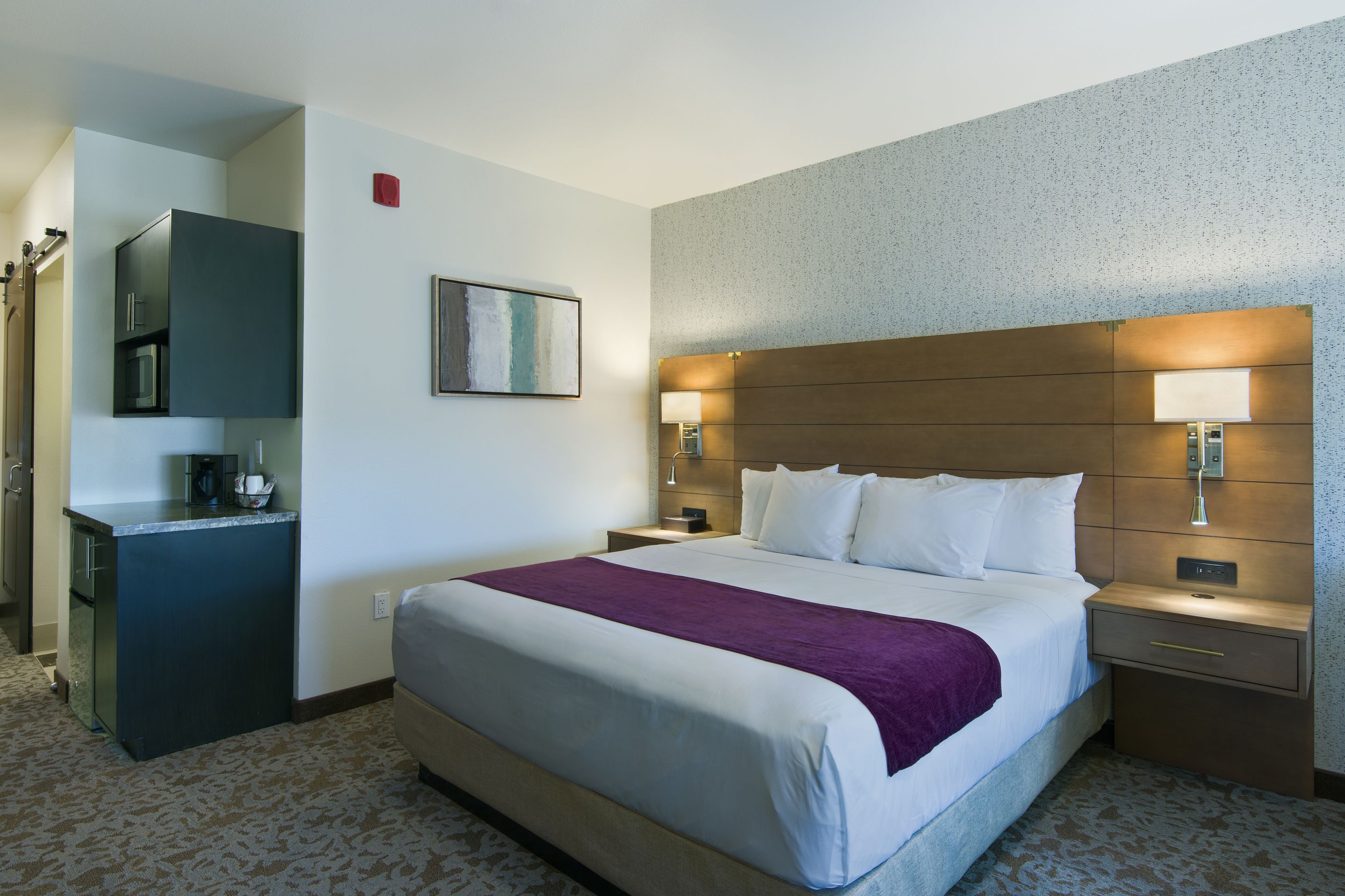 A hotel room with a large bed, a bedside table, a nightstand, a built-in desk with a microwave, and a wall-mounted TV.