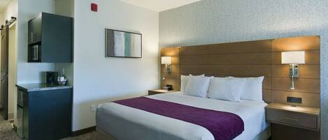 A hotel room with a large bed, a bedside table, a nightstand, a built-in desk with a microwave, and a wall-mounted TV.