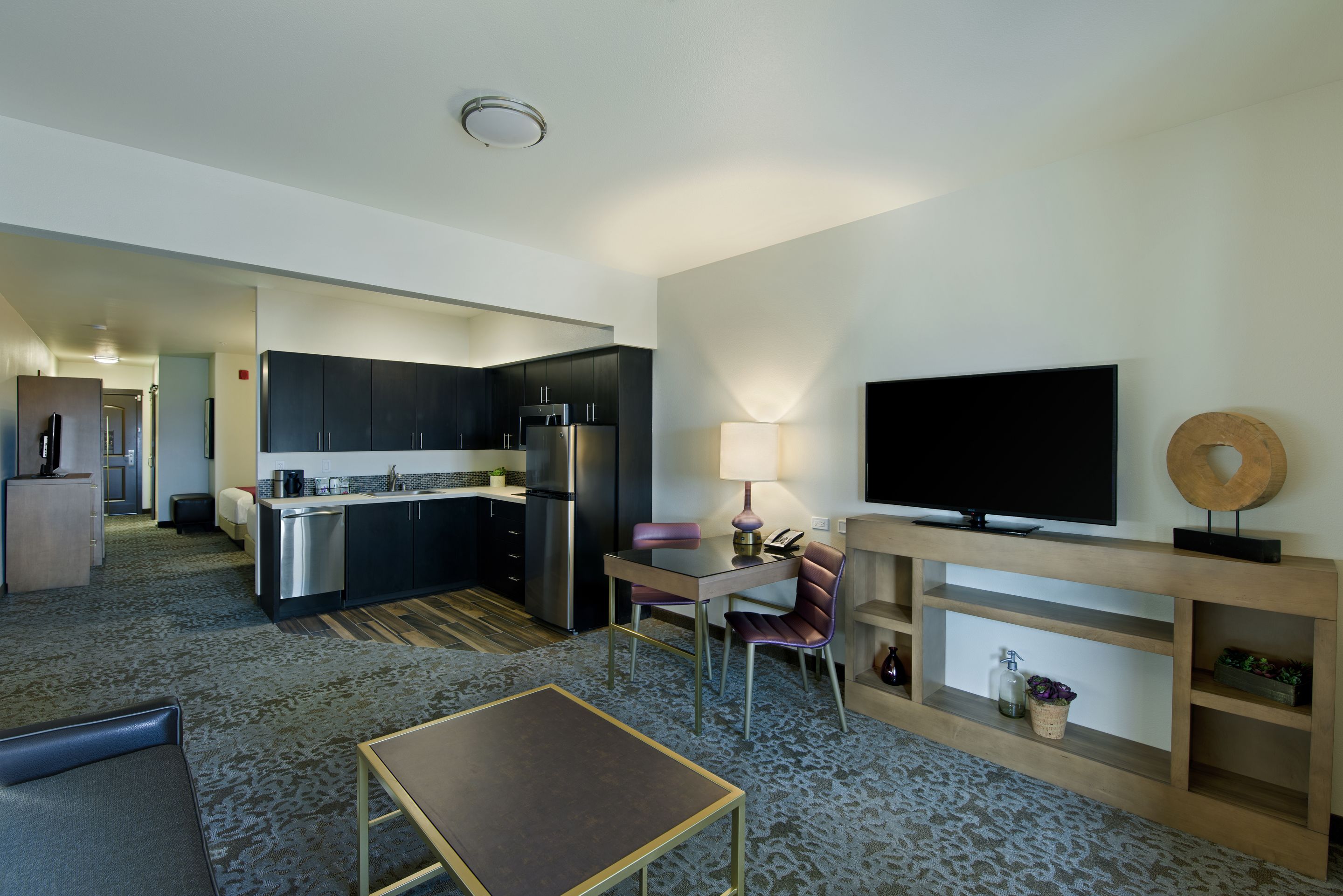 A modern hotel room with a kitchenette, a flat-screen TV, a dining area, and a living space.