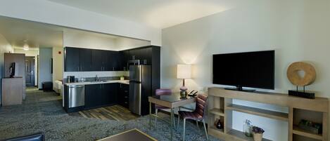 A modern hotel room with a kitchenette, a flat-screen TV, a dining area, and a living space.