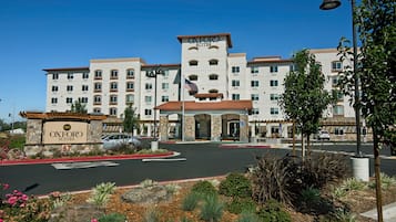 A multi-story hotel with a central entrance, a flag, and a landscaped area with plants and rocks.