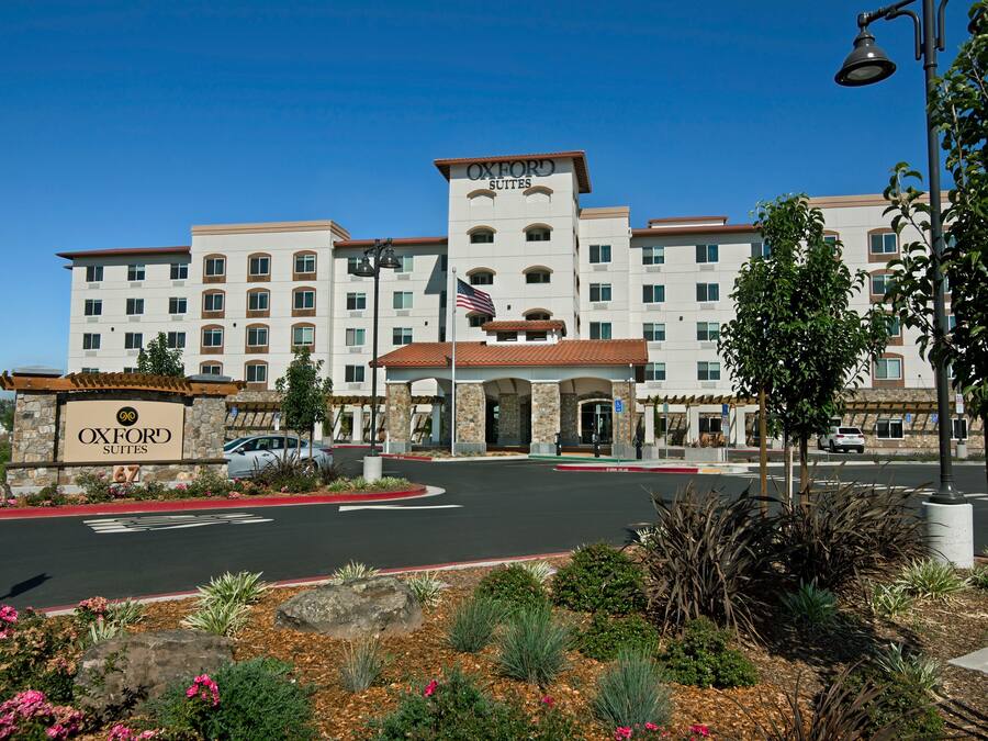 A multi-story hotel with a central entrance, a flag, and a landscaped area with plants and rocks.