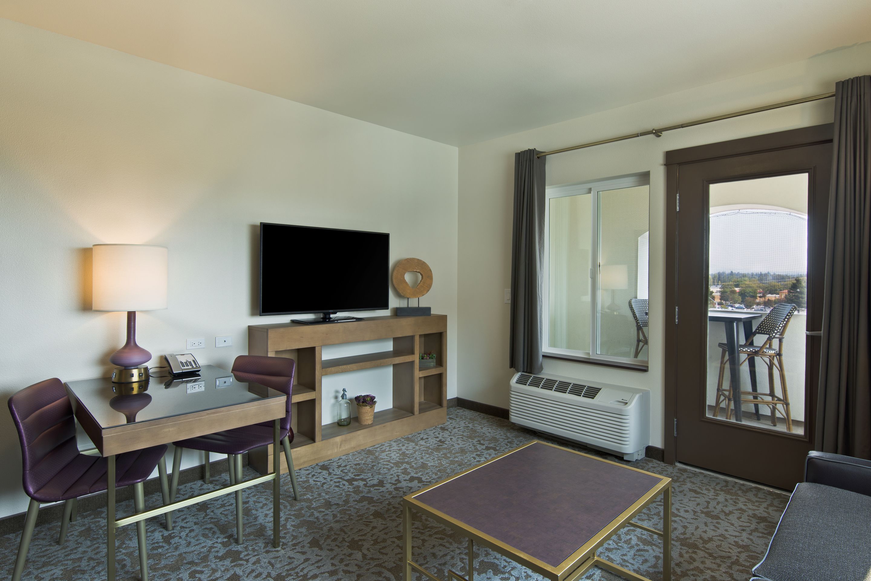 Habitación de hotel con televisor de pantalla plana, una mesa de comedor con sillas, una mesita pequeña, un sofá, un ventanal con vistas y un equipo de aire acondicionado.
