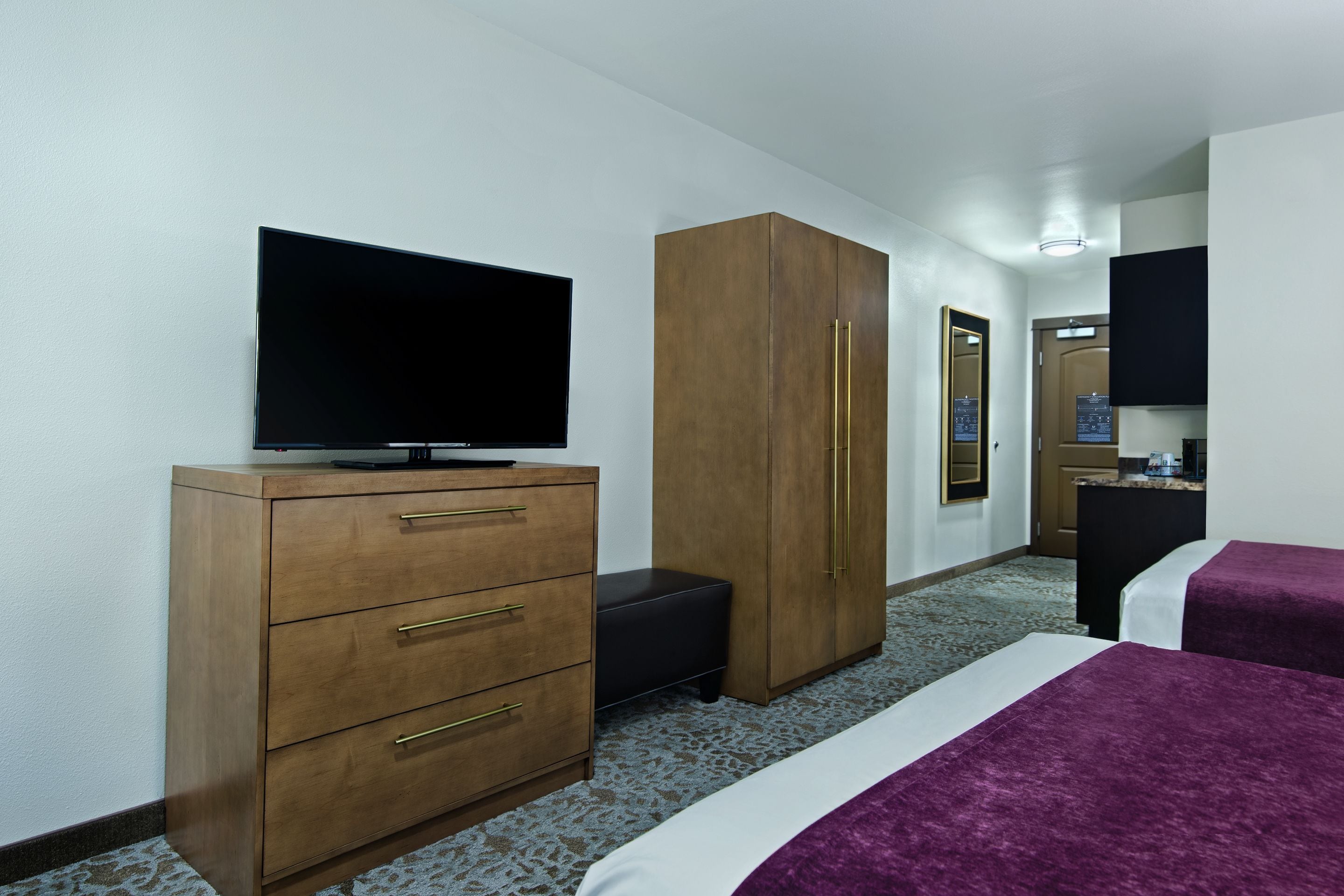 Quarto de hotel com televisão de tela plana sobre uma cômoda de madeira, um guarda-roupa, um banquinho preto pequeno e duas camas com roupa de cama roxa.
