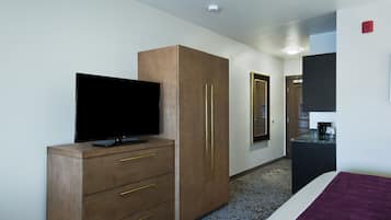 A hotel room with a flat-screen TV on a wooden dresser, a bed with a purple blanket, and a small kitchenette area.