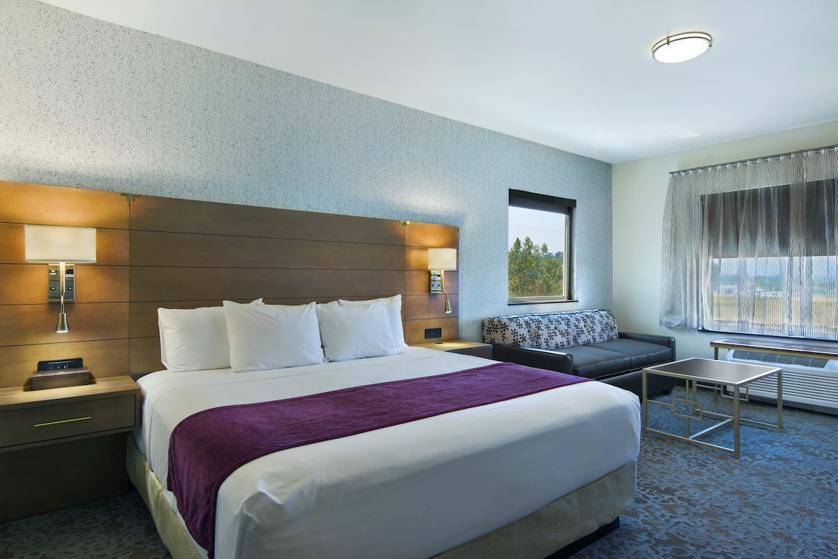 Ein Hotelzimmer mit einem großen Bett, einem Nachttisch mit Lampe, einer kleinen Couch und einem Fenster mit Vorhängen.