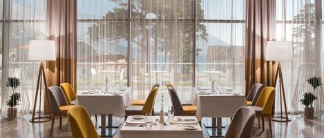 A dining area with tables set for two, large windows with sheer curtains, and modern chairs.