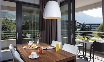 A dining area with a wooden table, white chairs, a white vase with flowers, a plate of croissants, a glass of orange juice, and a magazine.