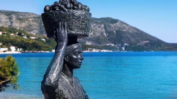 Uma estátua de uma pessoa carregando uma cesta na cabeça, com o mar azul e montanhas ao fundo.