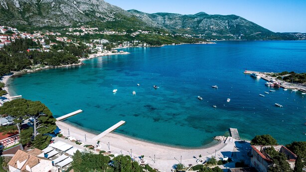 A coastal town with a marina, clear blue waters, and mountainous backdrop.