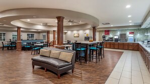 A spacious indoor area with a central seating area featuring a sofa and multiple tables and chairs. There is a kitchen area with a counter and bar stools. - Best Western Plus Galveston Suites (Galveston)