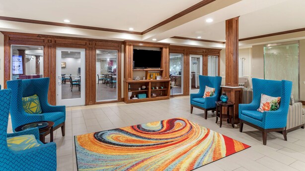 A spacious living area with blue armchairs, a colorful rug, wooden cabinets, and a television. - Best Western Plus Galveston Suites (Galveston)