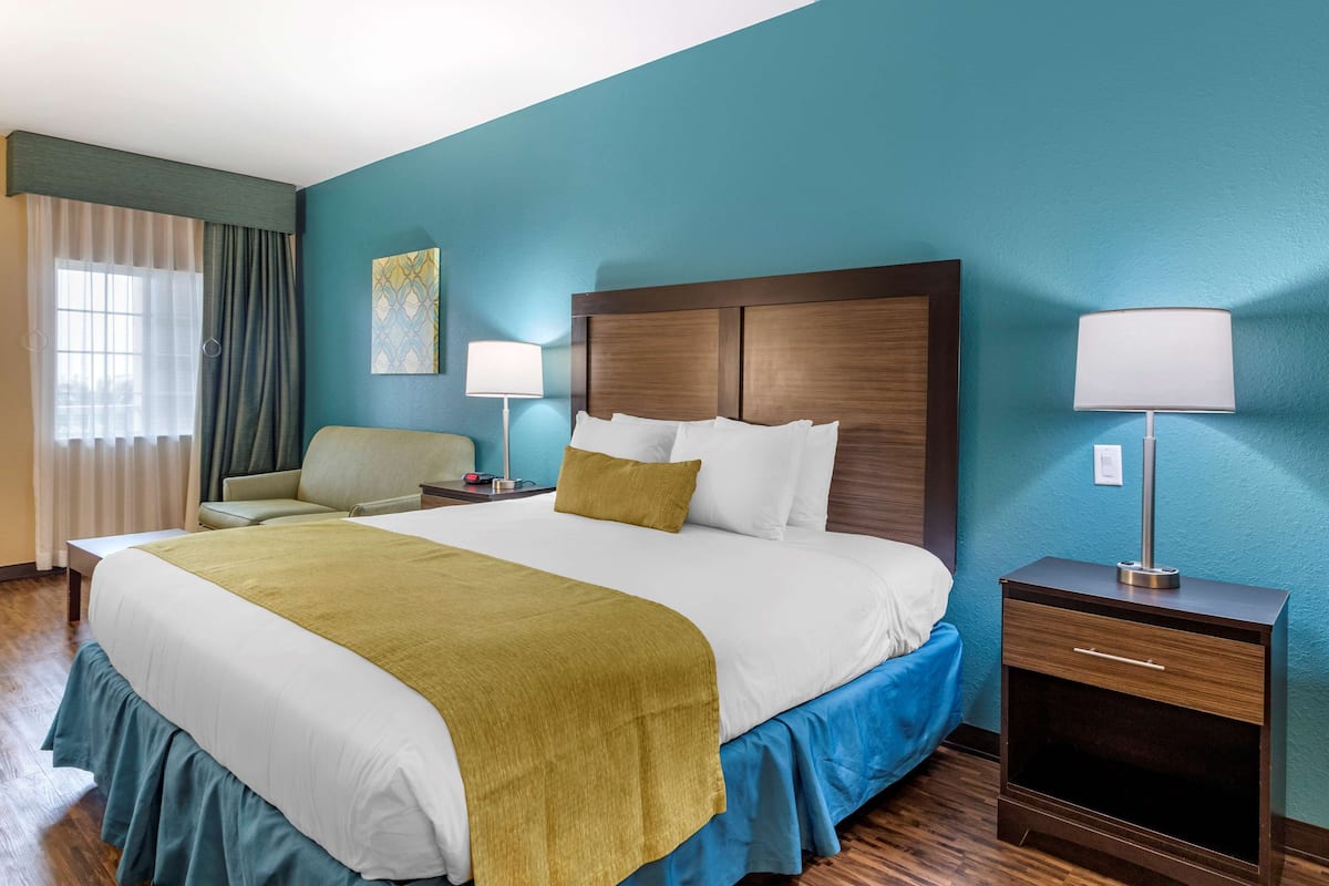 A hotel room with a large bed, a bedside table with a lamp, a chair, and a window with curtains.