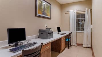 A small office space with two computer monitors, a printer, and a keyboard on a desk. There is a framed picture on the wall and a window with curtains.