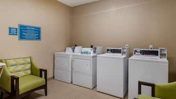 Un espace dédié à la buanderie, équipé d’une chaise, de machines à laver et d’un panneau indiquant les services de blanchisserie mis à la disposition des clients.