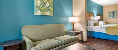 A hotel room with a green sofa, a wooden coffee table, a bedside table with a lamp, and a bed with a blue and white bedspread.