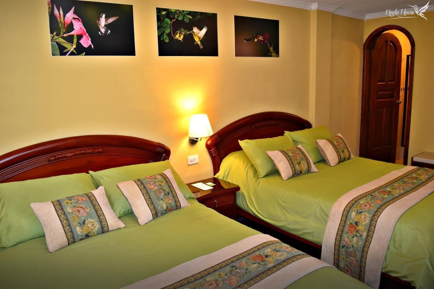 Ein Hotelzimmer mit zwei Betten, jedes mit grüner Bettwäsche und floralen Kissen. Dazwischen befindet sich ein Nachttisch mit einer Lampe. Die Wände sind gelb und es hängen drei gerahmte Bilder von Vögeln an der Wand.