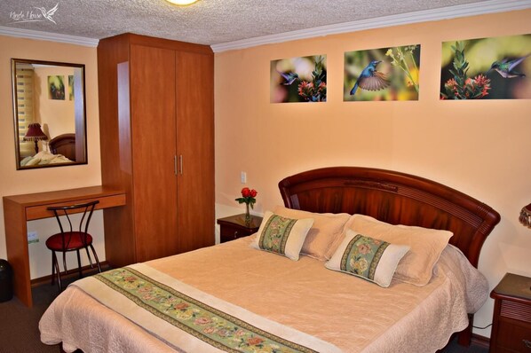 A bedroom with a large bed, a wooden wardrobe, a desk with a chair, and three framed pictures on the wall.