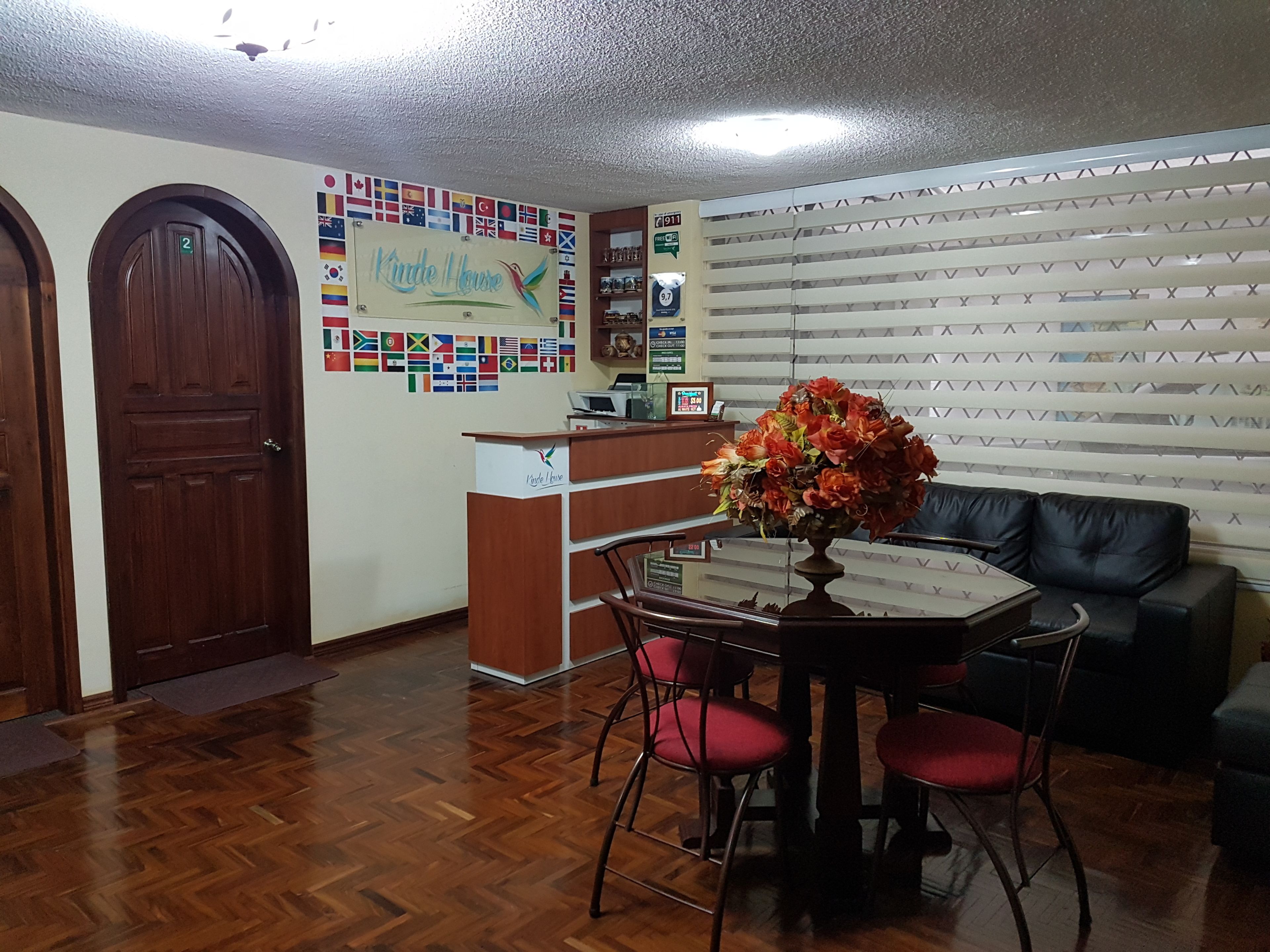 Un espace d’accueil comprenant un bureau en bois, un comptoir de réception, un mur décoré de drapeaux internationaux, un canapé en cuir noir et une table à manger avec des chaises rouges.