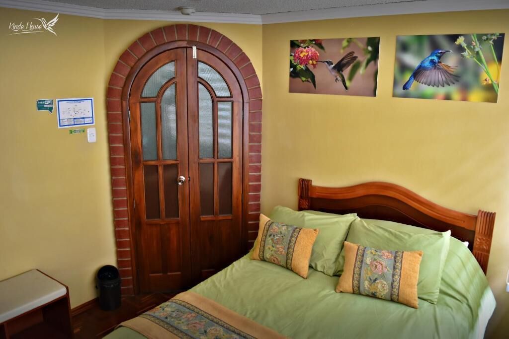 A bedroom with a wooden door, a bed with green bedding and floral pillows, and two framed bird pictures on the wall.
