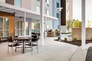 An outdoor patio area with tables and chairs, a stone wall, and a view of a building in the background. - Towneplace Suites Kansas City Airport (Kansas City)