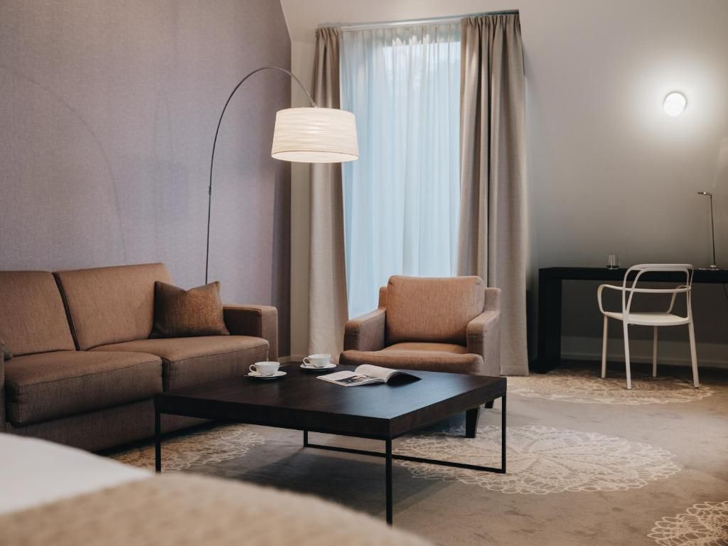 A modern hotel room with a sofa, armchair, coffee table, and a desk with a chair.