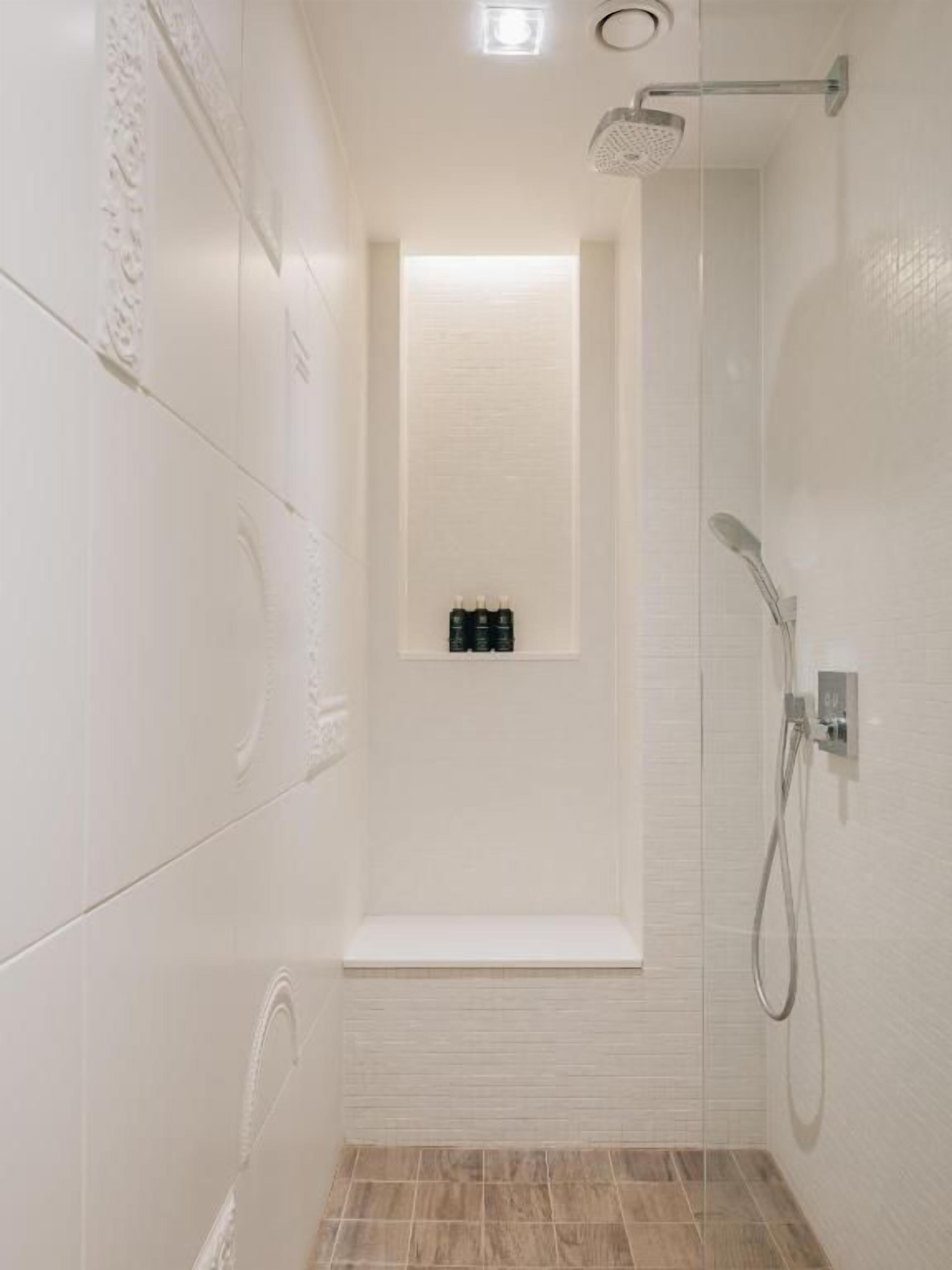 A narrow shower area with a bench, a wall-mounted shelf with toiletries, and a showerhead.