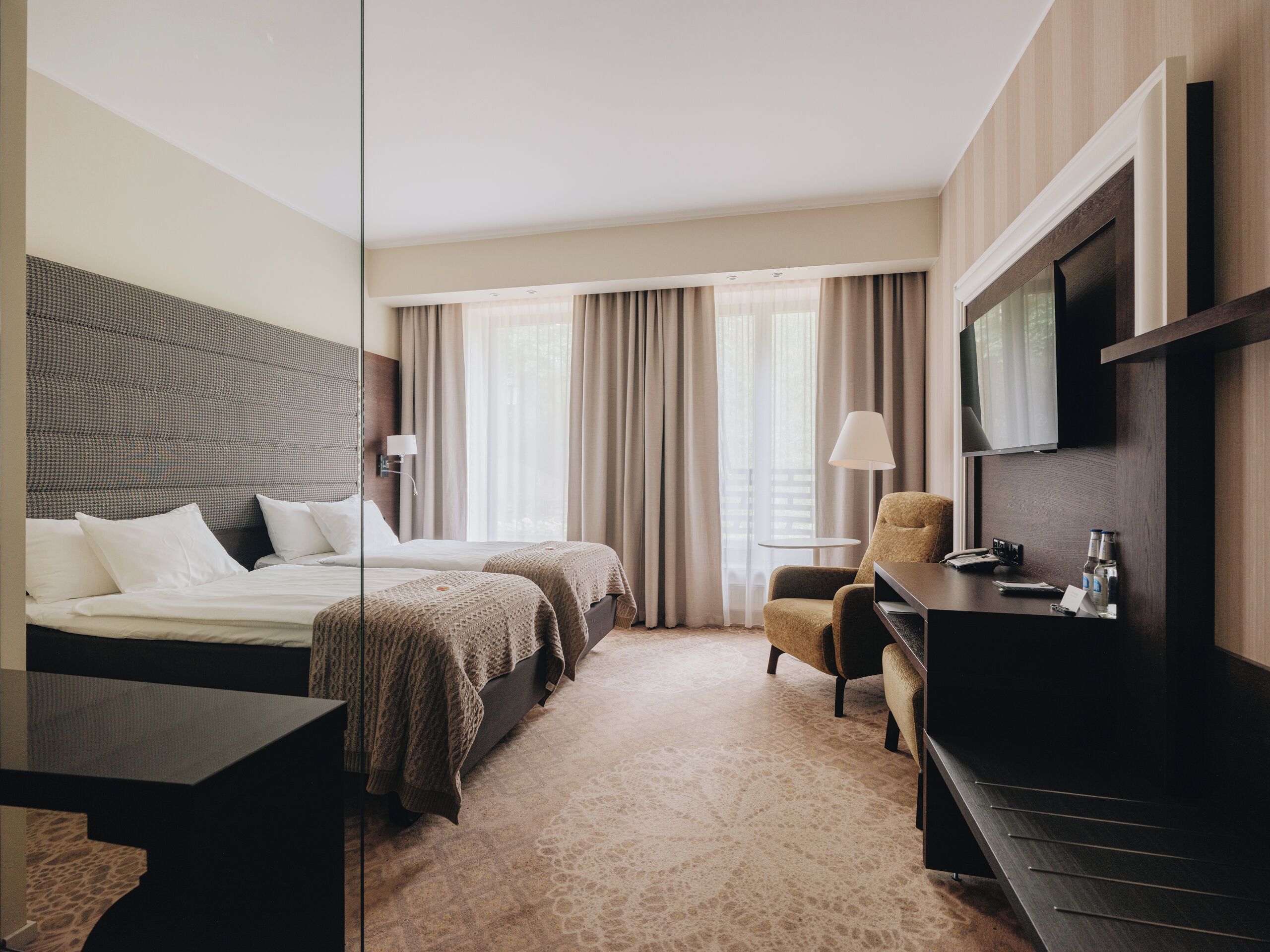 Habitación de hotel con cama, dos mesitas de noche, una silla, escritorio, televisor y ventana con cortinas.