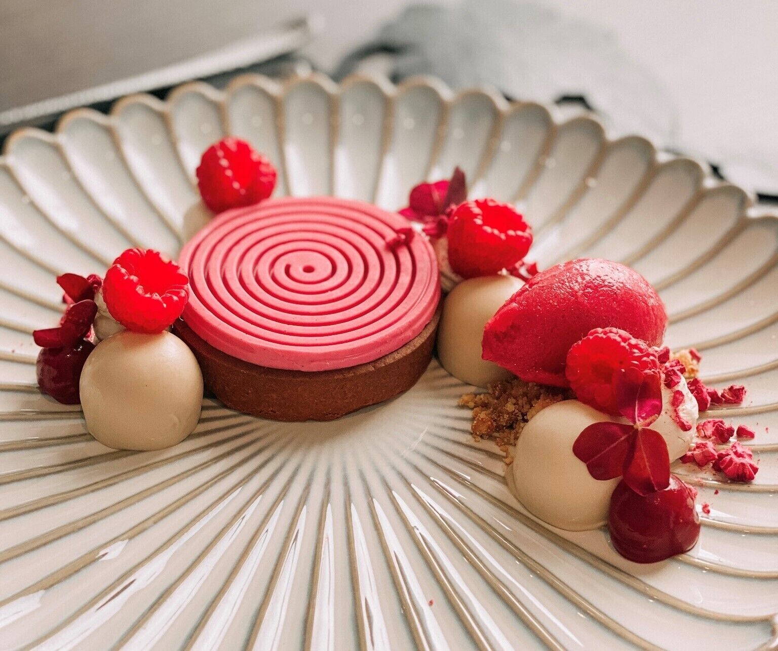 A dessert with a spiral design on a plate, surrounded by small round sweets and raspberry pieces.