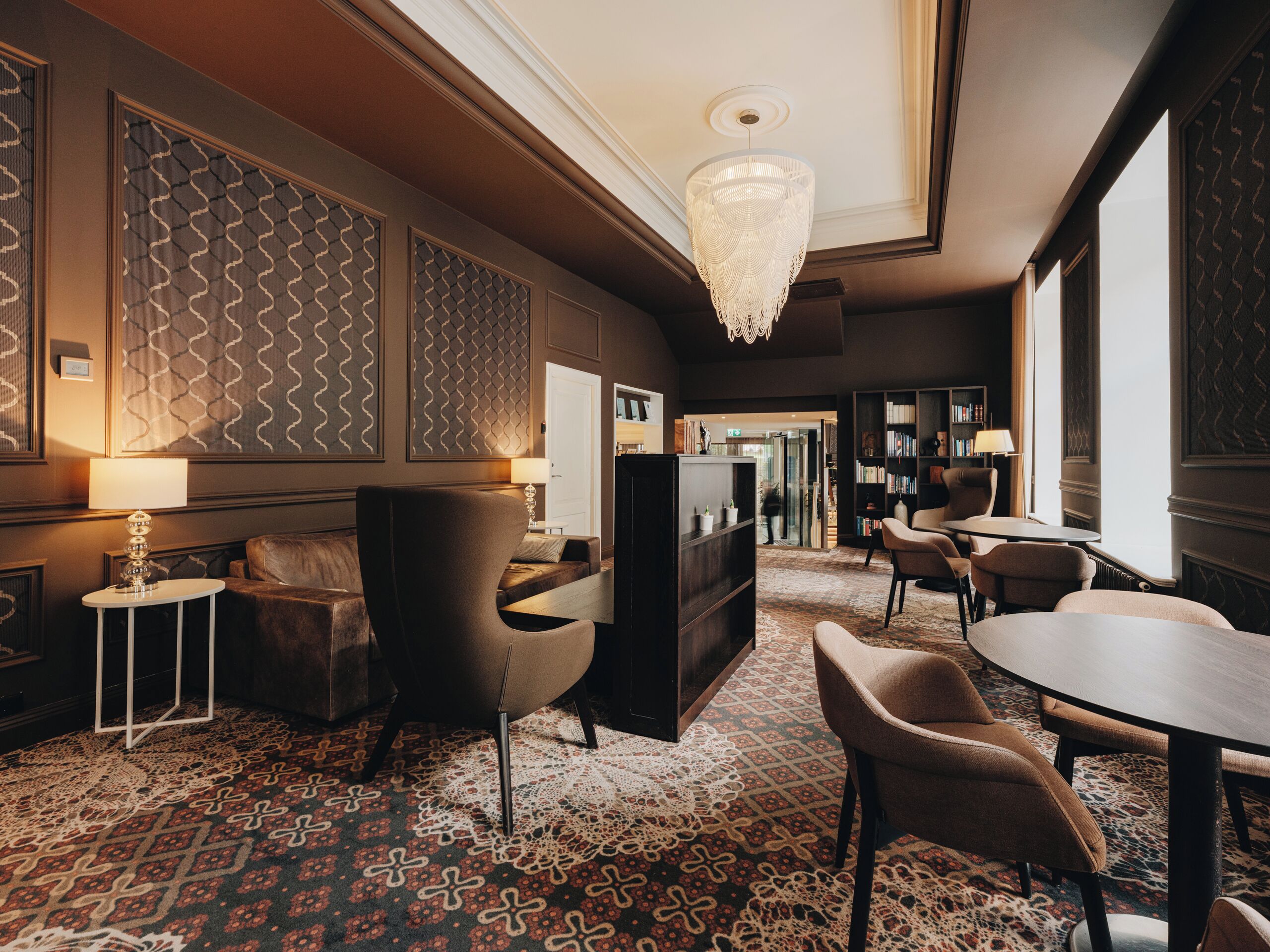 A hotel lobby with a patterned carpet, a chandelier, seating areas, and a bookshelf.