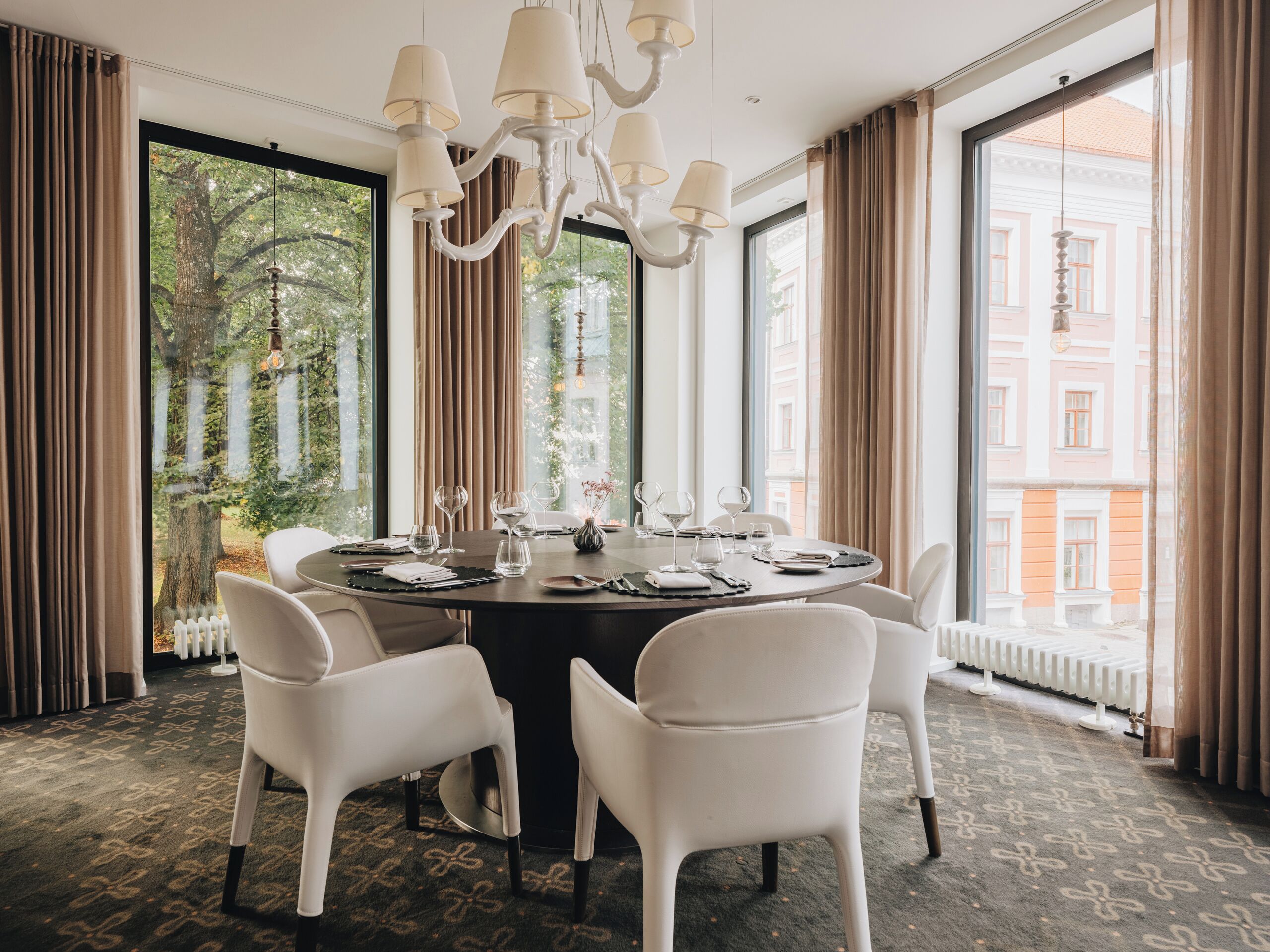 A dining area with a round table set for six, surrounded by white chairs, with a chandelier above and a view of a building through large windows.