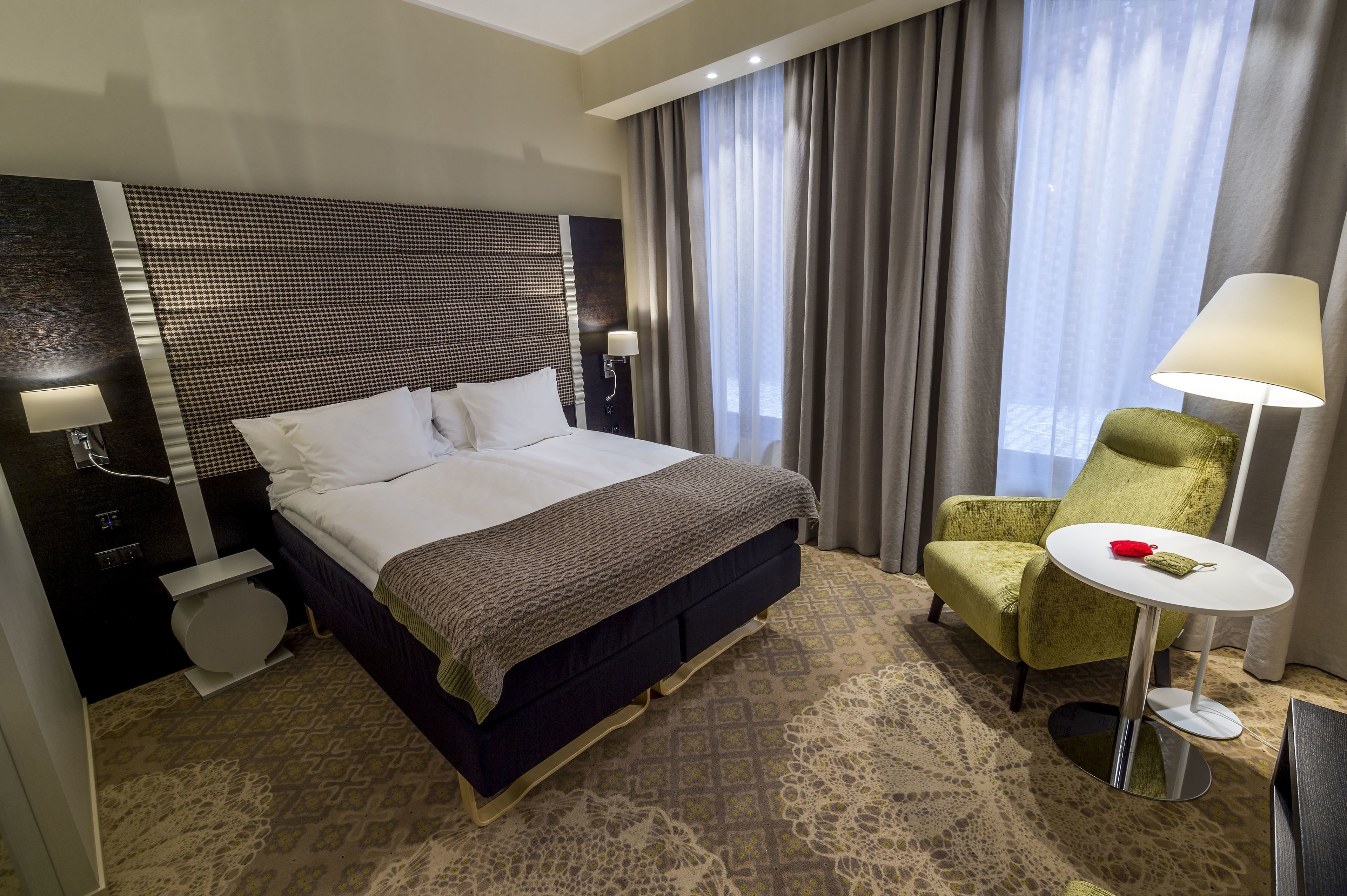 A hotel room with a large bed, a green armchair, a small round table, and a nightstand with a lamp.