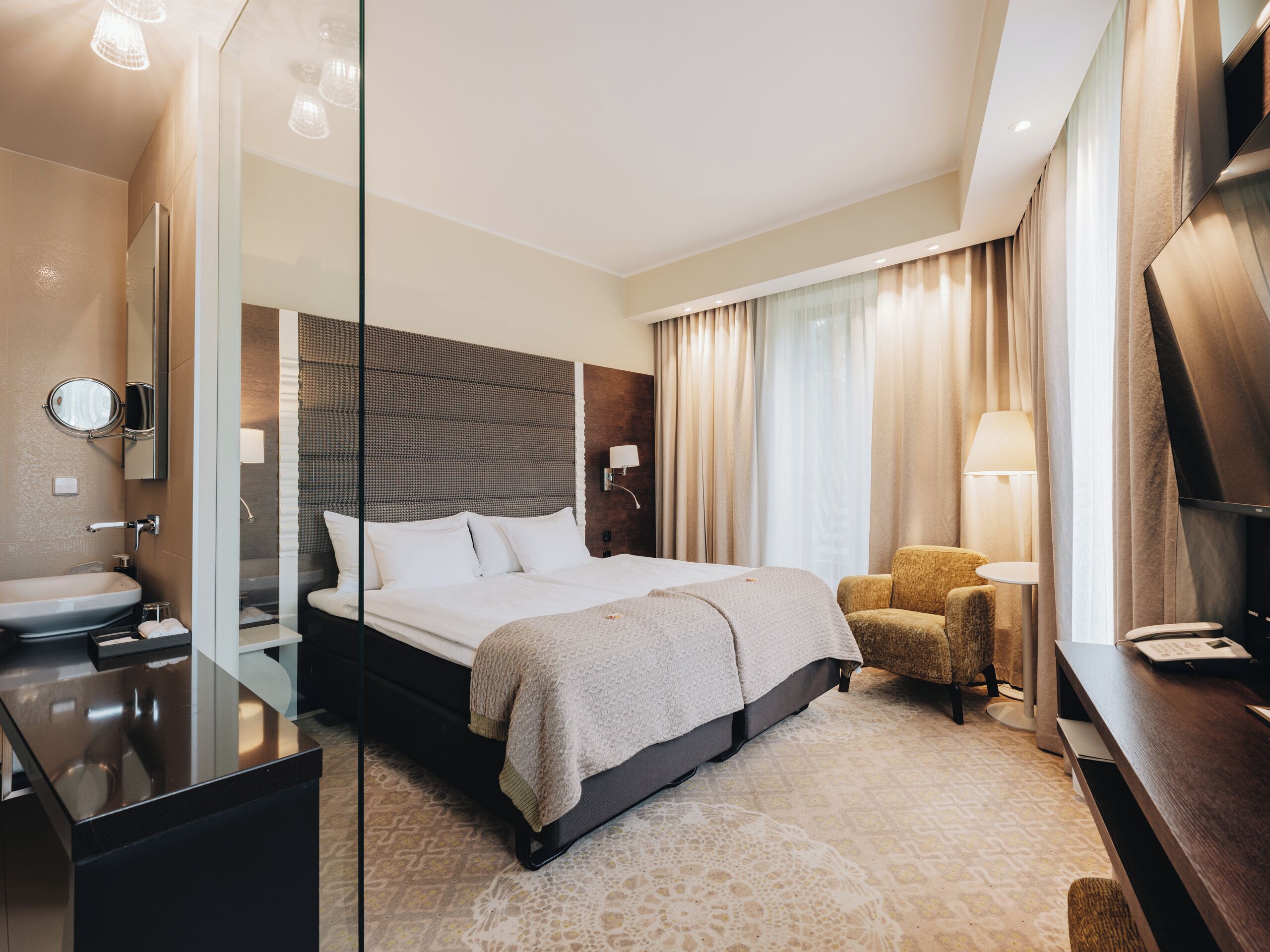 Ein modernes Hotelzimmer mit einem großen Bett, einem Nachttisch, einem Sessel, einem Schreibtisch und einem Badezimmer mit Waschbecken und Spiegel.