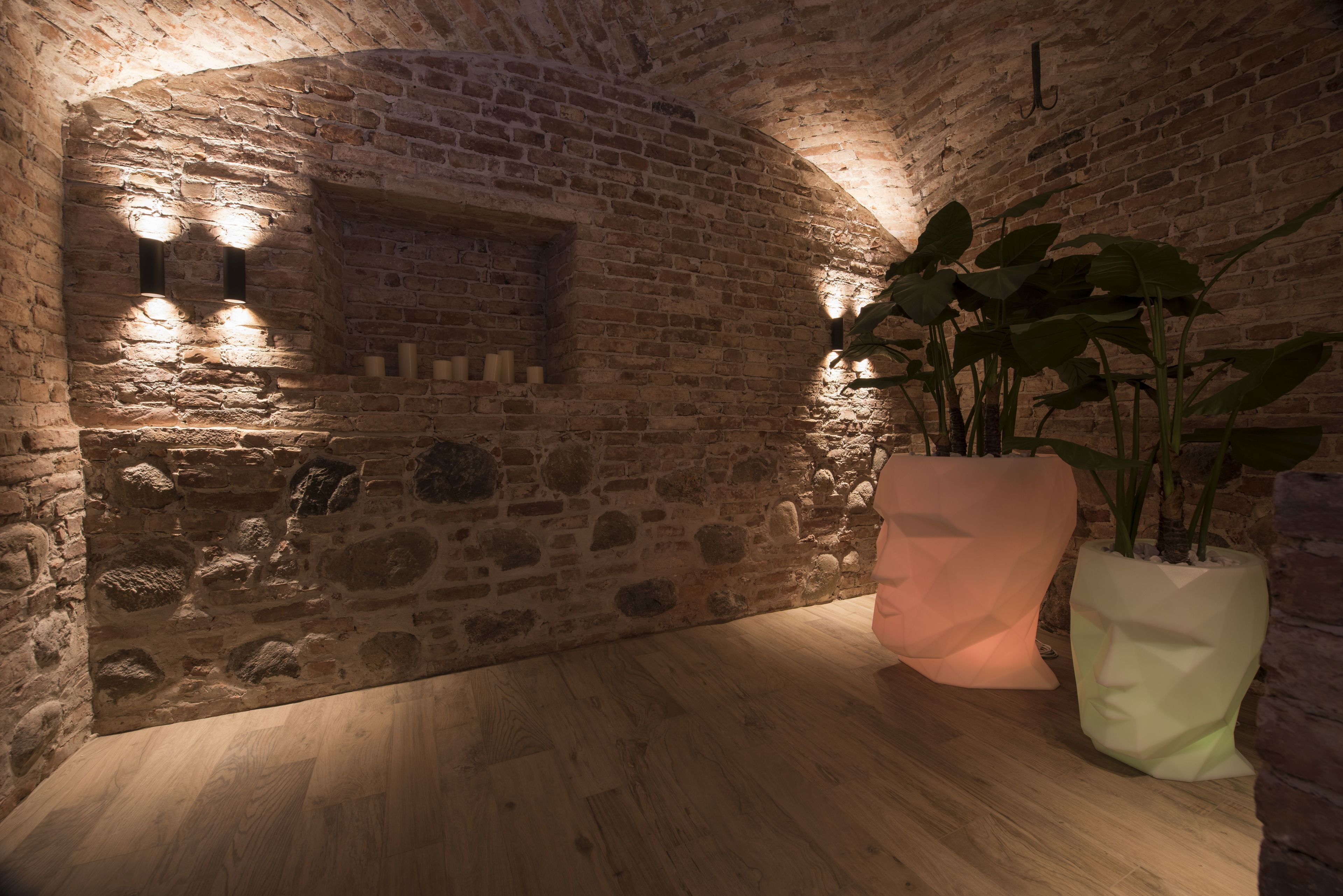 A stone-walled room with wooden flooring, two wall-mounted lights, and large potted plants.