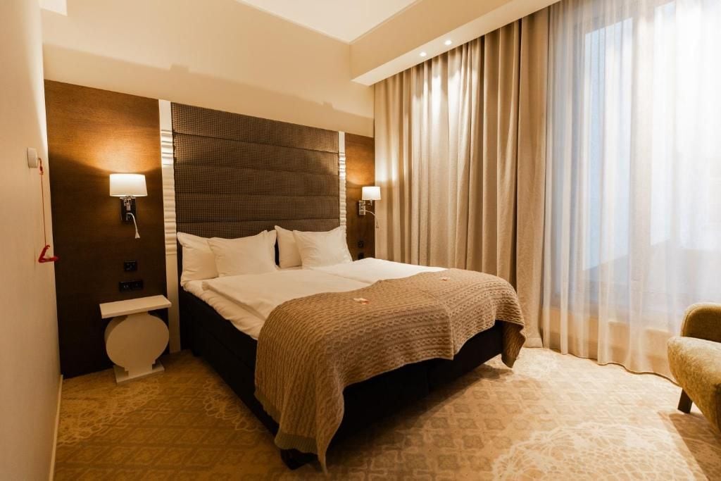 A hotel room with a large bed, a bedside table, a lamp, and a window with curtains.