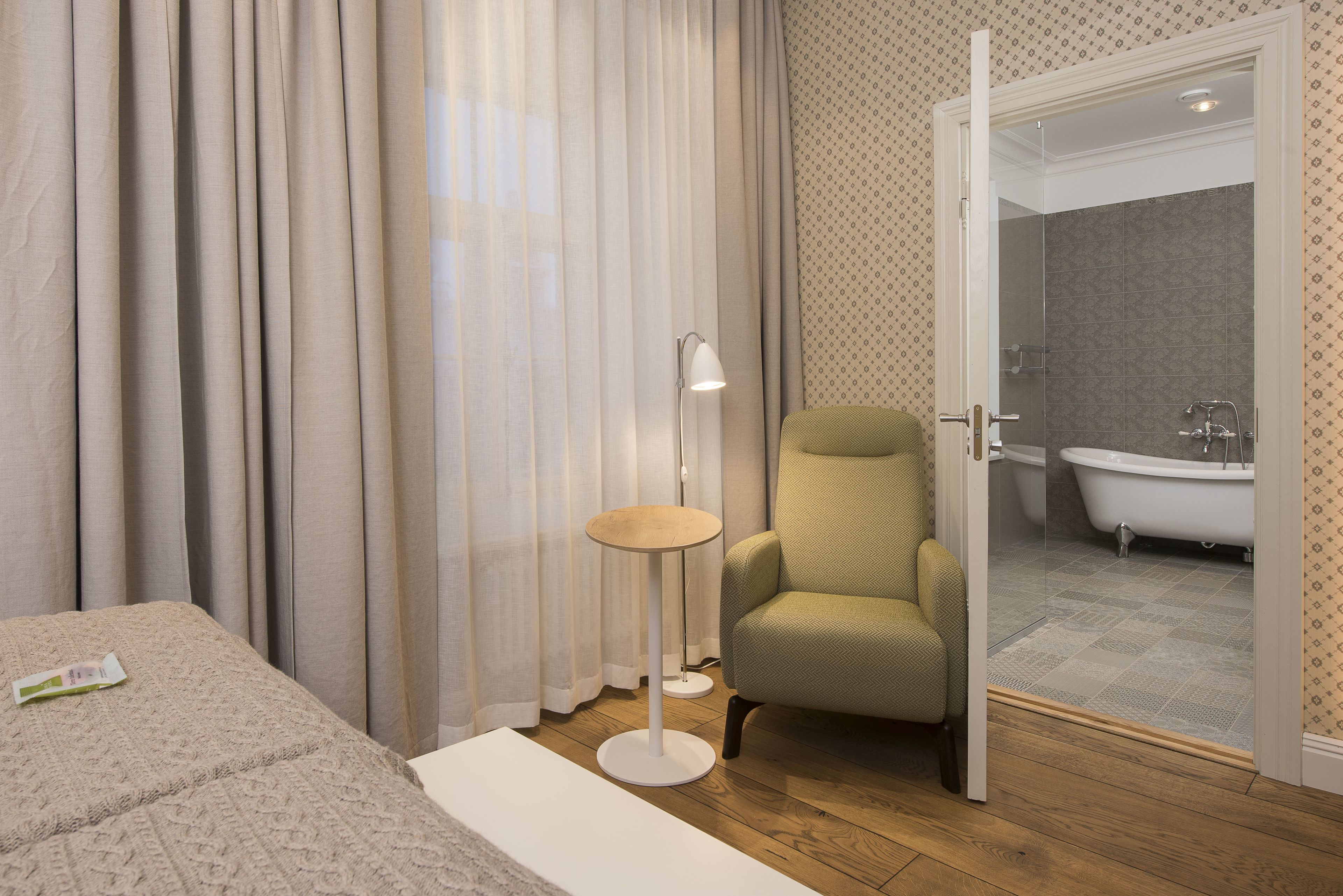 Ein Hotelzimmer mit einem Bett, einem grünen Sessel, einer Stehlampe und einem durch eine offene Tür einsehbaren Badezimmer.