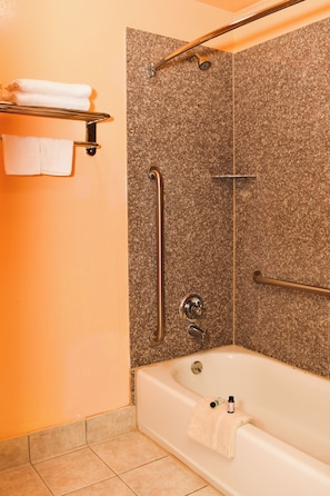 A bathroom with a bathtub, grab bars, a shelf with towels, and a shower area.