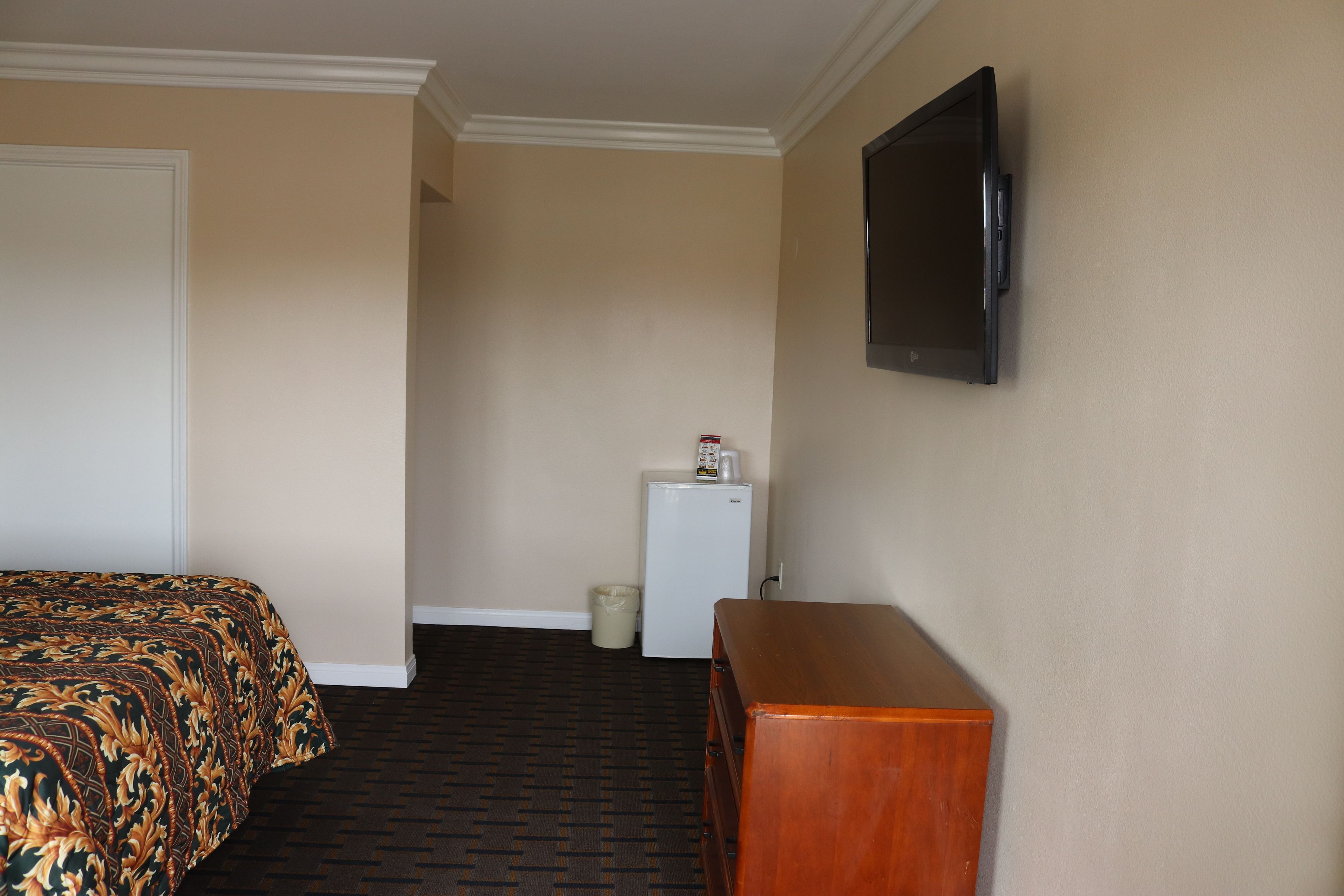 Ett hotellrum med en säng, en byrå, en tv som är monterad på väggen och ett litet kylskåp.