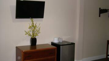 A room with a wooden dresser, a black refrigerator, a flat-screen TV mounted on the wall, and a vase with yellow flowers on the dresser.