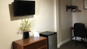 A hotel room with a wooden dresser, a black refrigerator, a flat-screen TV mounted on the wall, a small table with a vase of flowers, and a black office chair. - Fullerton Lodge (Fullerton)