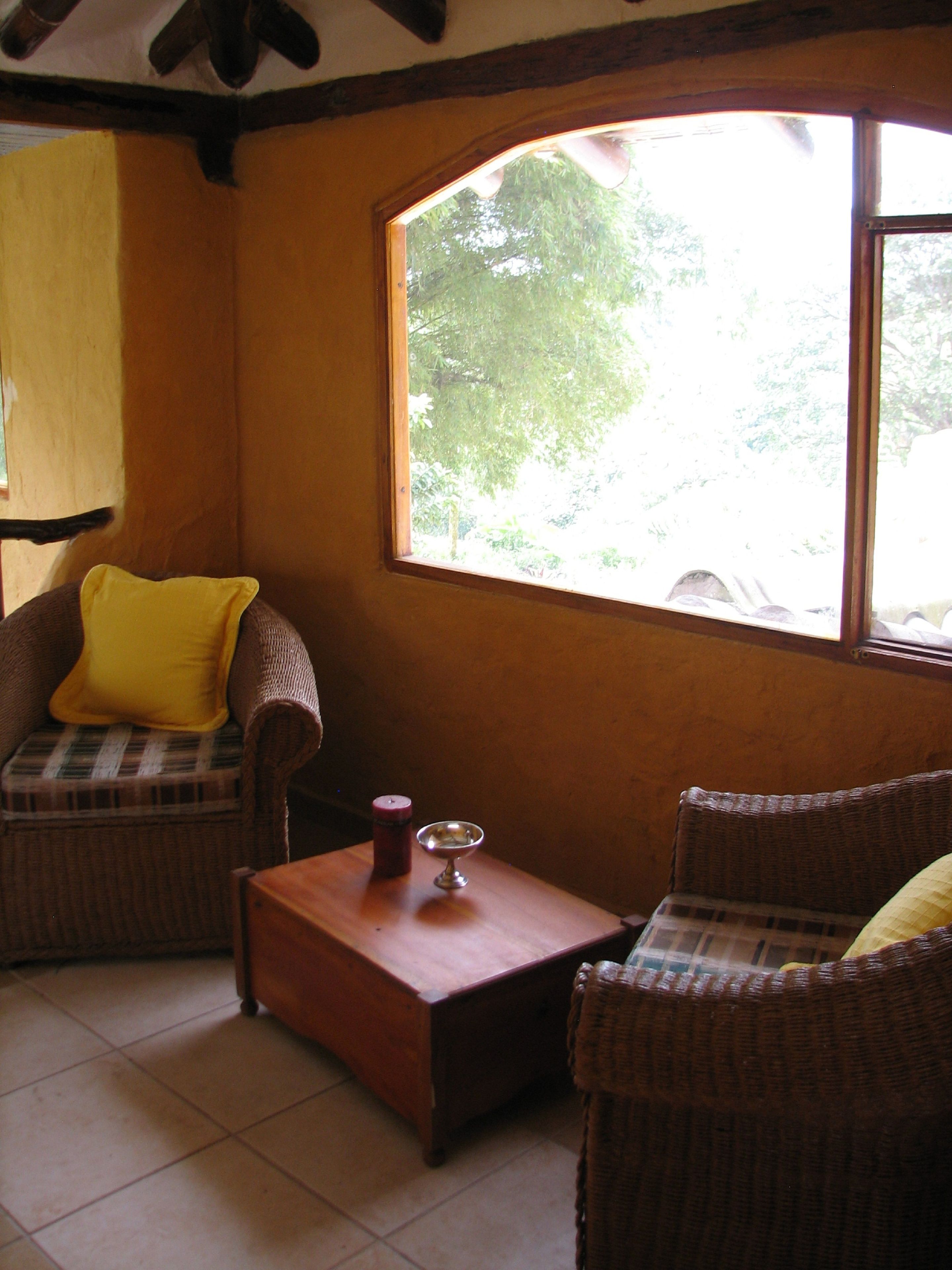 A cozy room with wicker furniture, a wooden coffee table, and a large window offering a view of greenery outside.