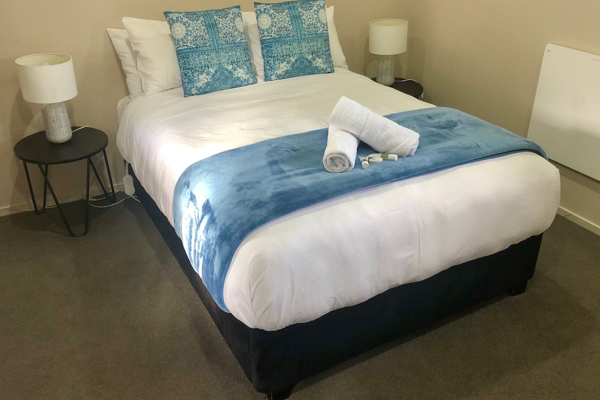 A neatly made bed with a blue and white bedspread, two matching pillows, and a rolled white towel on the bed. There are two bedside tables, each with a lamp and a small decorative item. The room has a carpeted floor and plain walls.