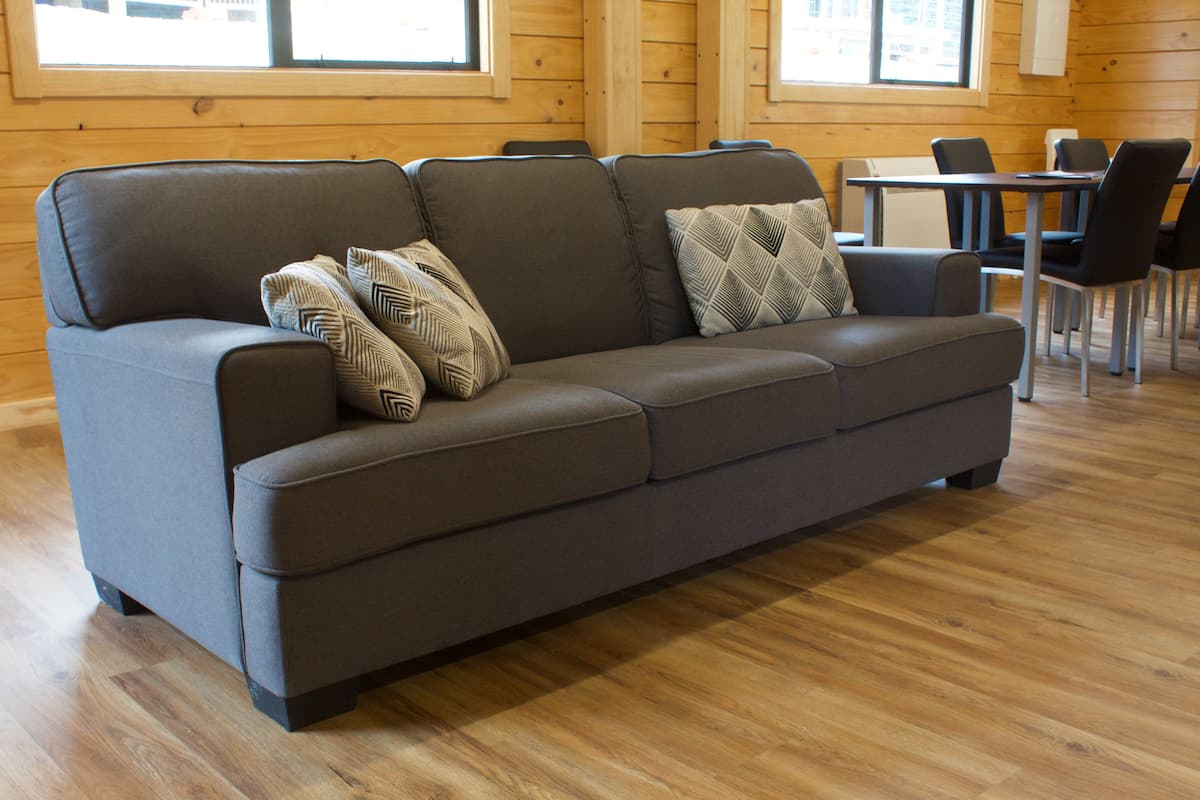 A modern log cabin interior with a grey sofa, patterned pillows, and a dining area with chairs and a table.