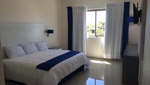 A hotel room with a large bed, a nightstand, a wall-mounted TV, and a window with curtains. - MH Grand Hotel (Ometepec)